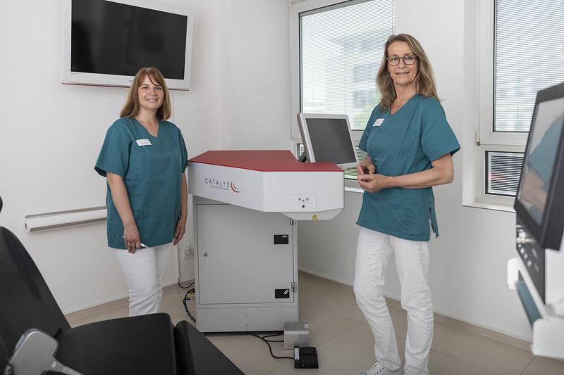 Zwei Frauen in blaugrünen Kitteln stehen neben den medizinischen Geräten und demonstrieren in einem hellen, modernen Klinikraum starken Teamgeist.