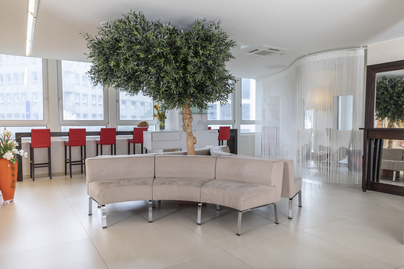 Moderne Lounge mit beigefarbenem, geschwungenem Sofa, roten Barhockern und Platz für Teambesprechungen unter einem großen künstlichen Baum.