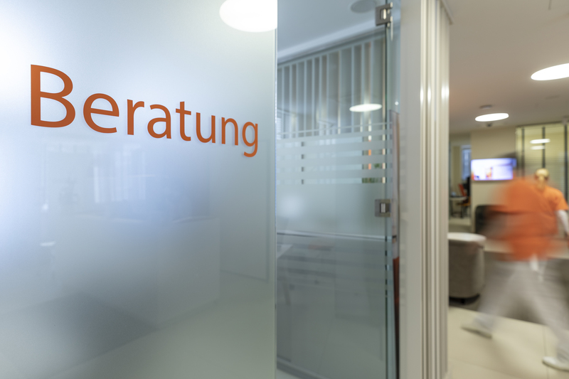 Milchglastür mit der Aufschrift "Beratung"; unscharfes Büro, Teammitglied im Hintergrund sichtbar.
