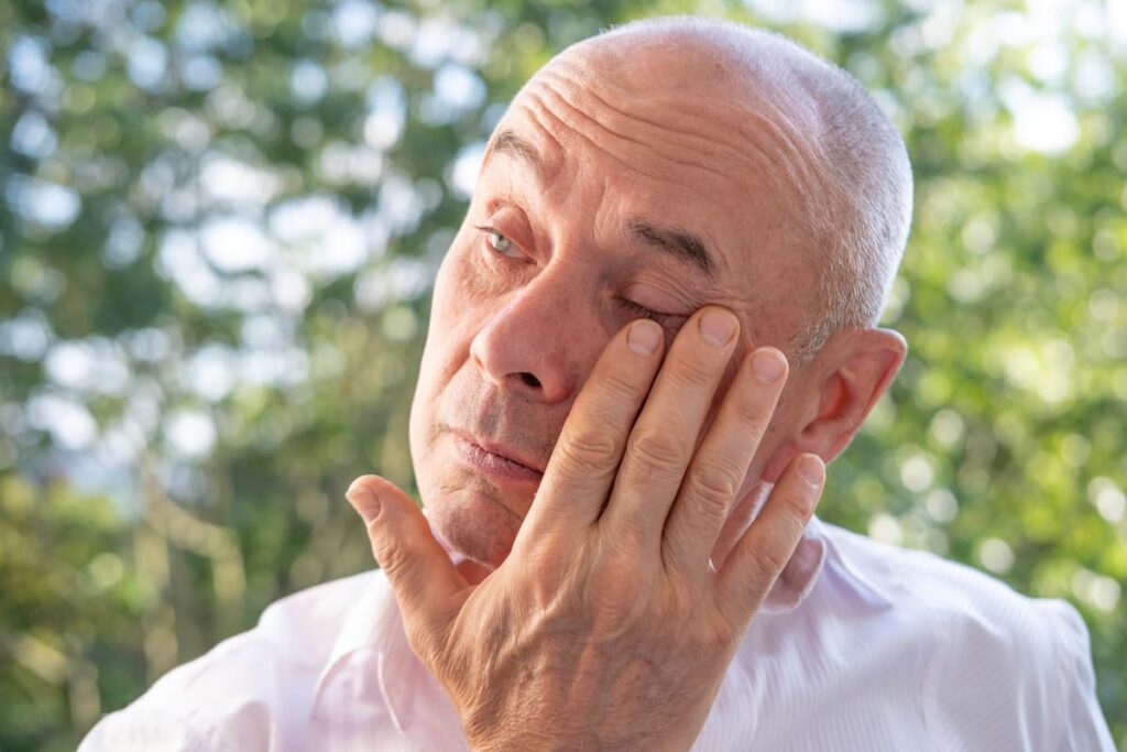 Ein glatzköpfiger Mann im weißen Hemd reibt sich im Freien das Auge, möglicherweise auf der Suche nach Erleichterung vor der Minimalinvasiven Glaukomchirurgie (MIGS).