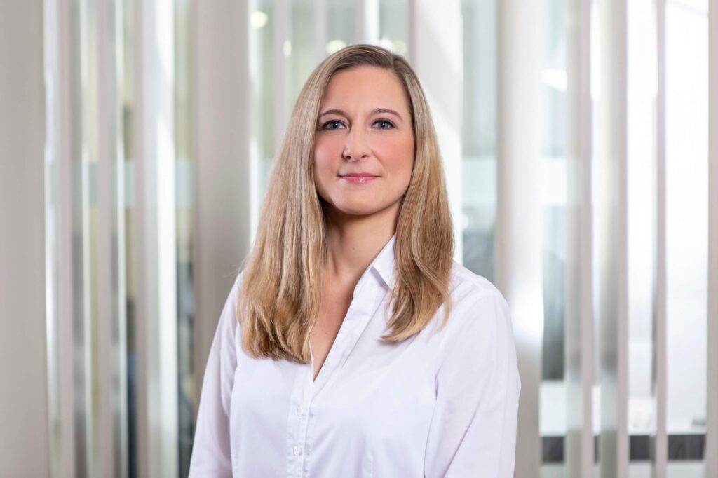 Frau mit langen blonden Haaren und weißem Hemd steht in einem Innenraum vor einer Glasscheibe und gehört zu einem Team.