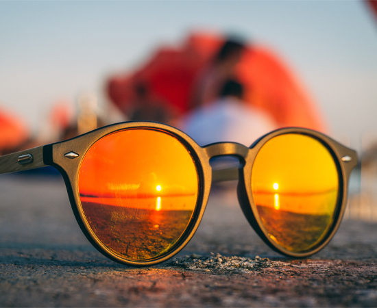 Die runde Sonnenbrille mit orangefarbenen Gläsern reflektiert einen Sonnenuntergang - perfekt für alle, die auf die Erkrankung AMD achten.