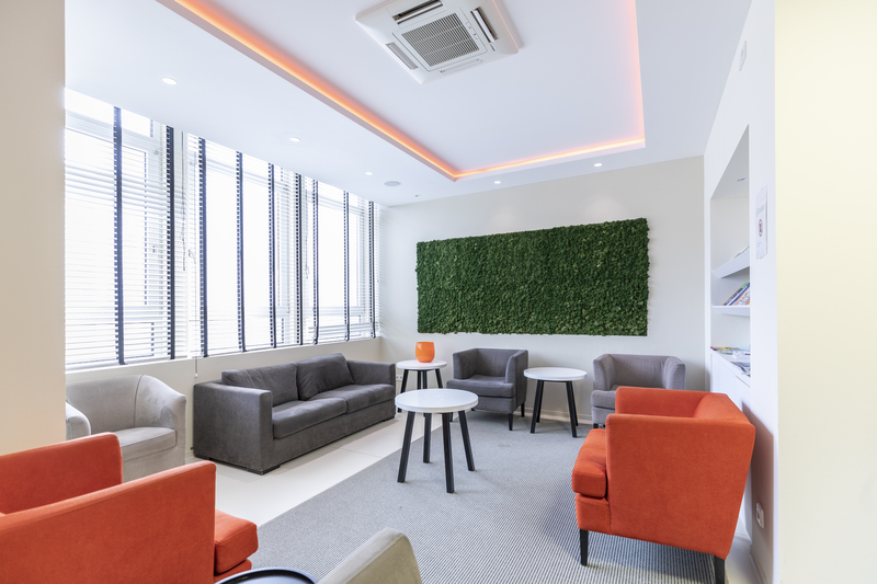 Moderne Lounge mit orangefarbenen und grauen Stühlen, kleinen Tischen, großen Fenstern und einer grünen Pflanzenwand für Feedbackgespräche.
