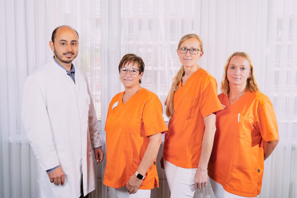 Ein Arzt in einem weißen Kittel steht neben drei Krankenschwestern in orangefarbenen Kitteln und gibt in einem hellen Raum Feedback.